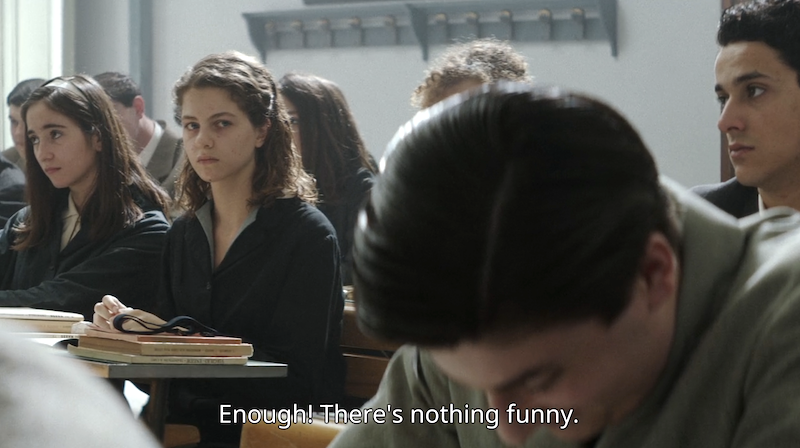 Gino laughs at Alfonso in class while Lenù looks at him with disappointment. The caption on the photo is a line of dialogue from the teacher, saying "Enough! There's nothing funny."