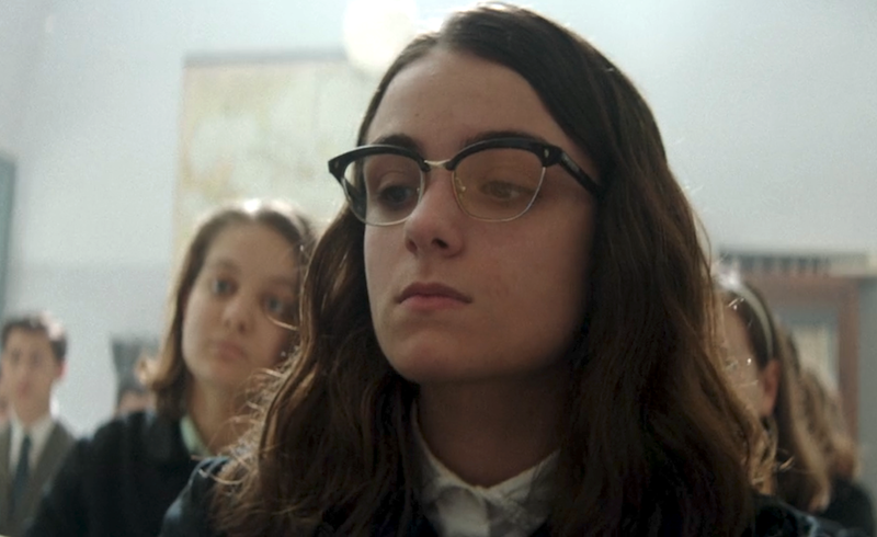 A girl with glasses looks at Lenù with disdain as she walks in front of the class to retrieve her essay from the teacher.