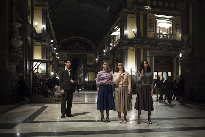 Pasquale stands off to the side and Lenù, Carmela, and Lila stand together with ice cream at the Galleria Principe.